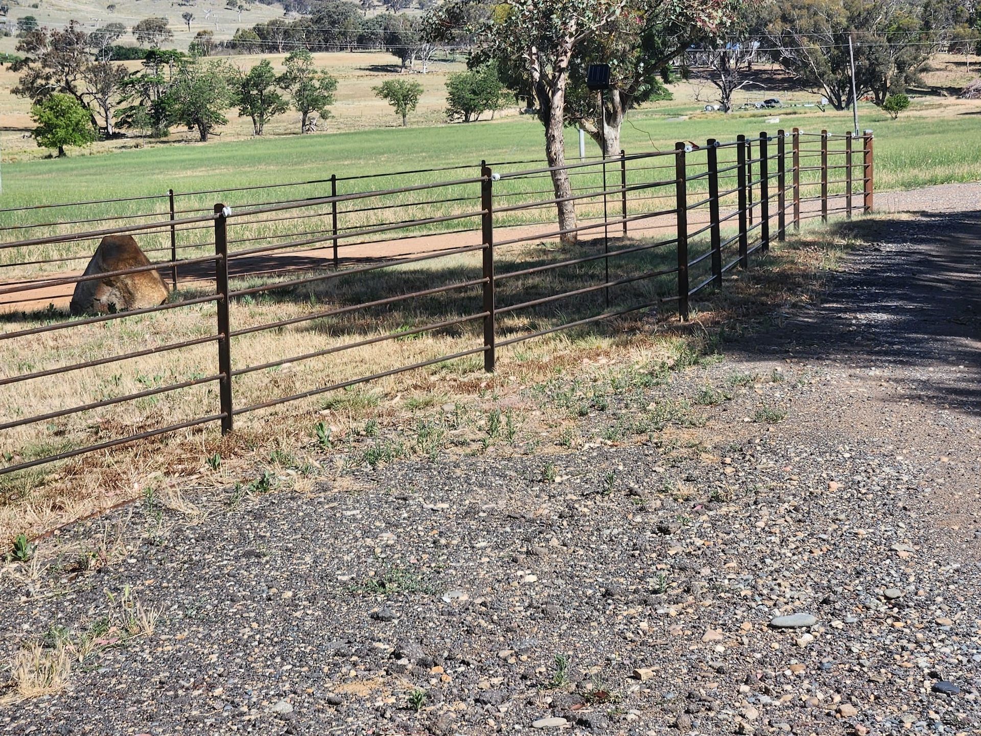 Metal fence bordering a gravel road and grassy field with trees under a blue sky.