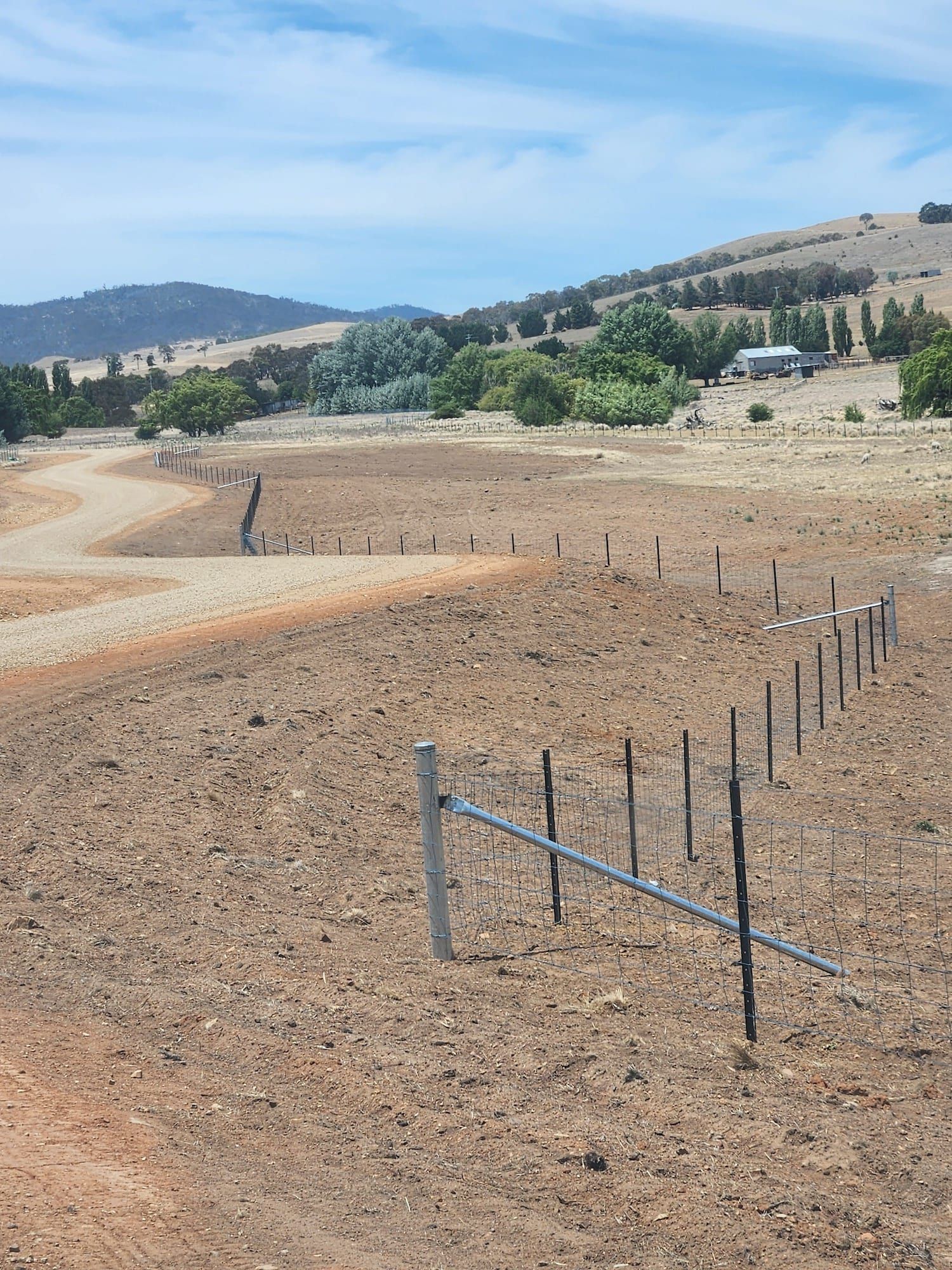 Barren field with fence, dirt road leading to trees and a building on a hillside under a partly cloudy sky.