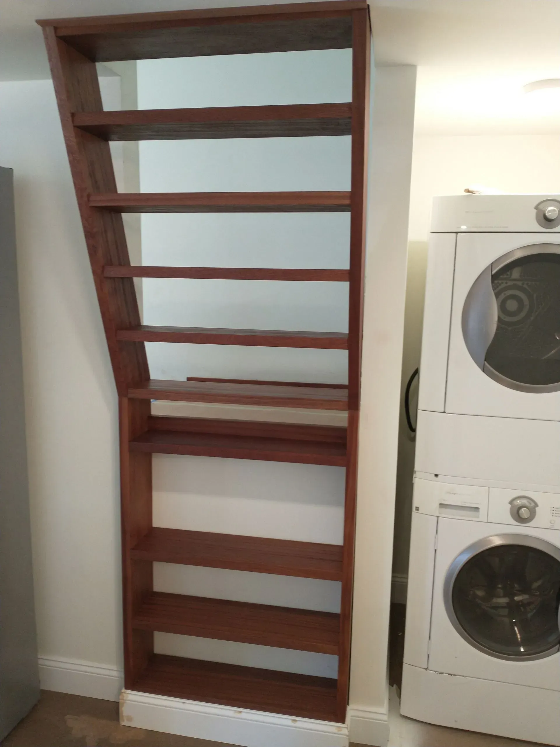 Tall, slanted wooden bookshelf against a white wall; washer and dryer in the background.