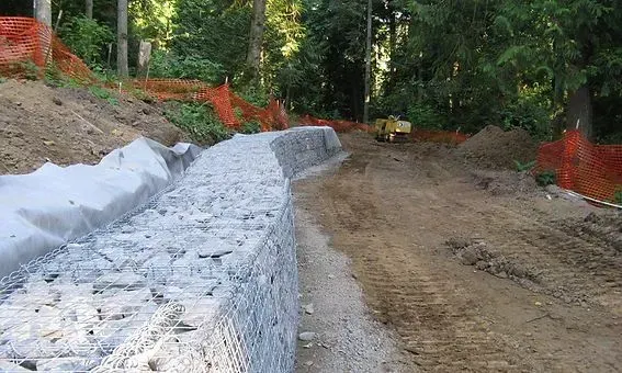 Construction site with a stone retaining wall along a dirt path, bordered by safety fencing and trees.