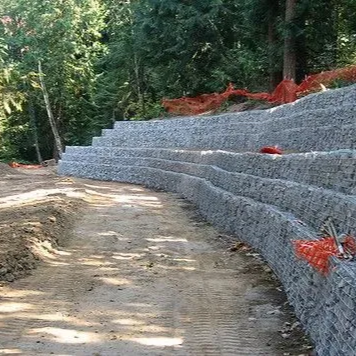 Gabion retaining wall constructed along a dirt path in a wooded area. Orange safety fencing visible.