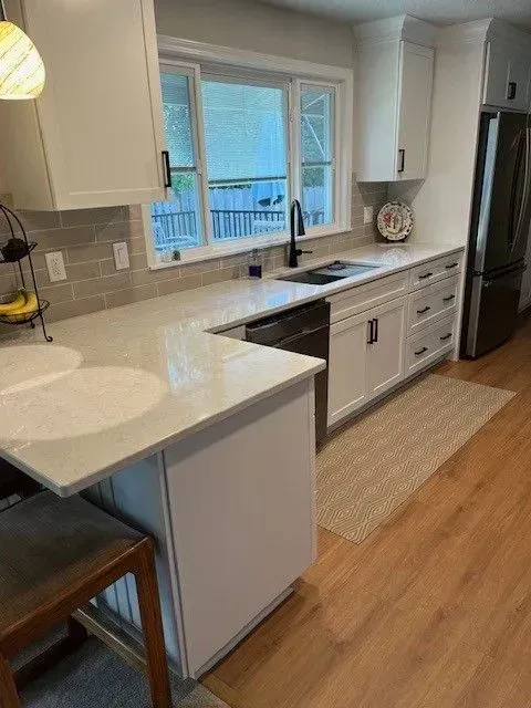Modern white kitchen with countertops, cabinets, black faucet, and refrigerator. Wooden floor and rug.