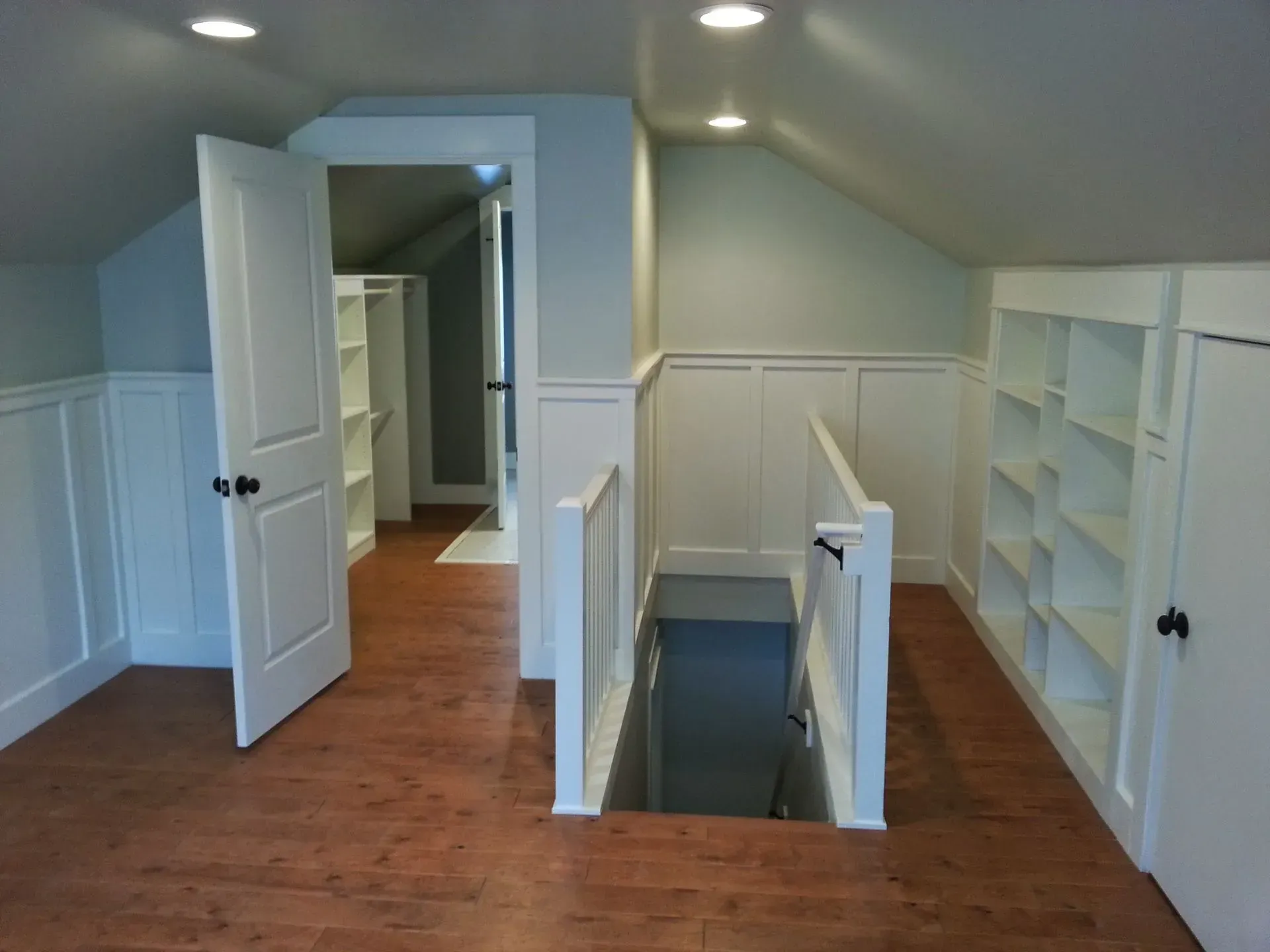 Attic room with wood floor, white bookshelves, and a staircase leading to a dark opening in the floor.