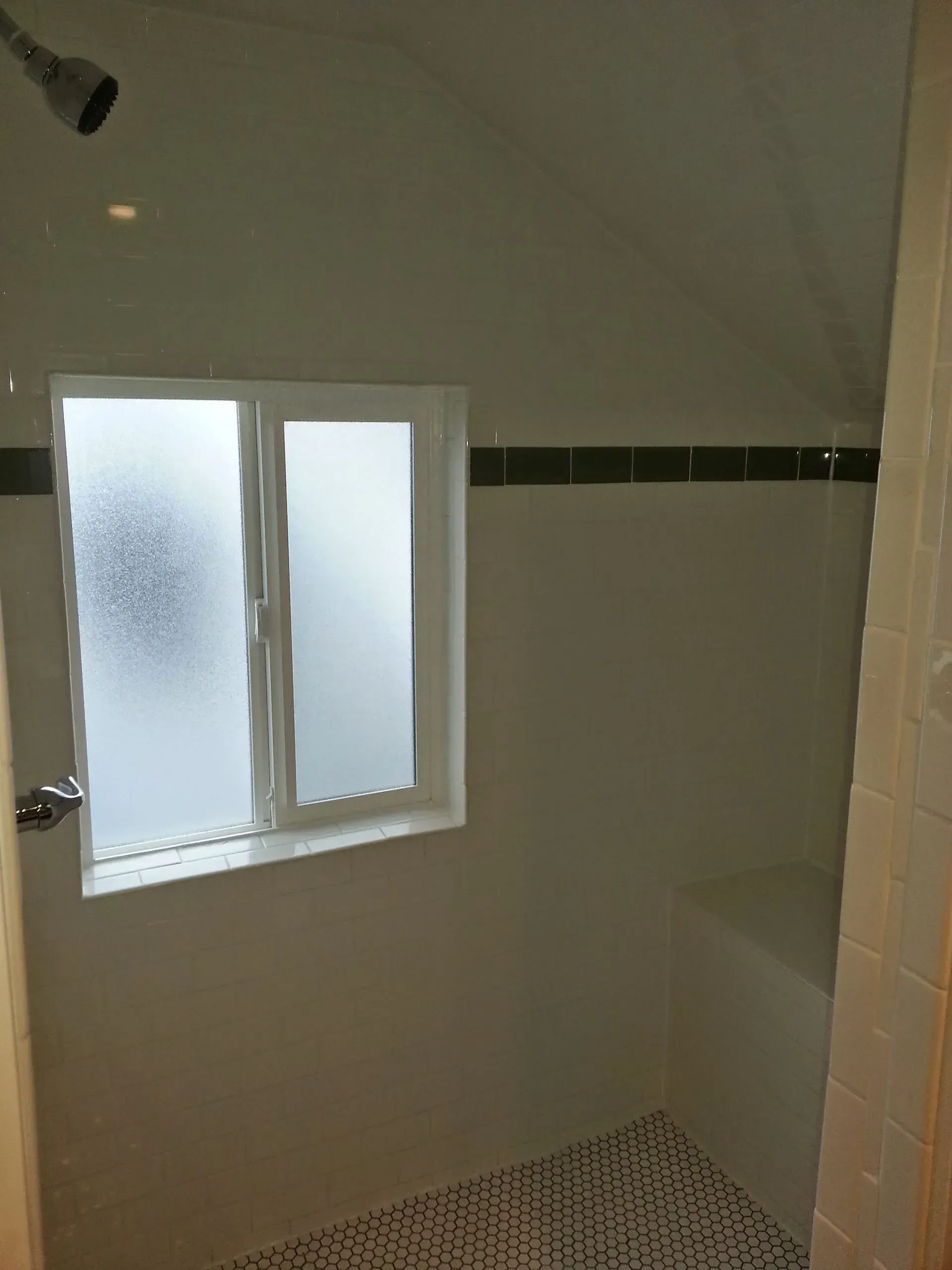 Bathroom with frosted window, showerhead, white walls, black tile accents, and mosaic tile floor.