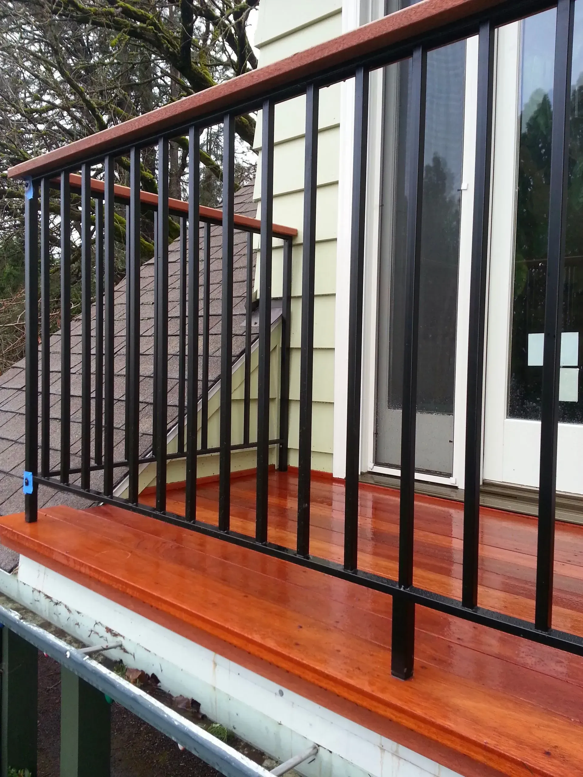 Black metal railing with wooden top and base, on a porch with reddish-brown wood flooring.