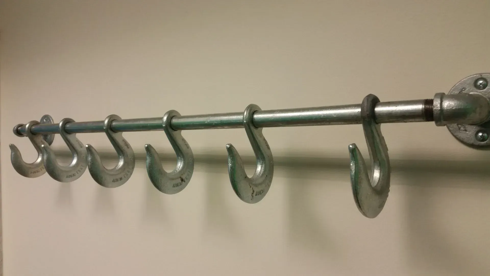 Metal hook rack mounted on a beige wall, with seven large S-shaped hooks hanging from a silver pipe.