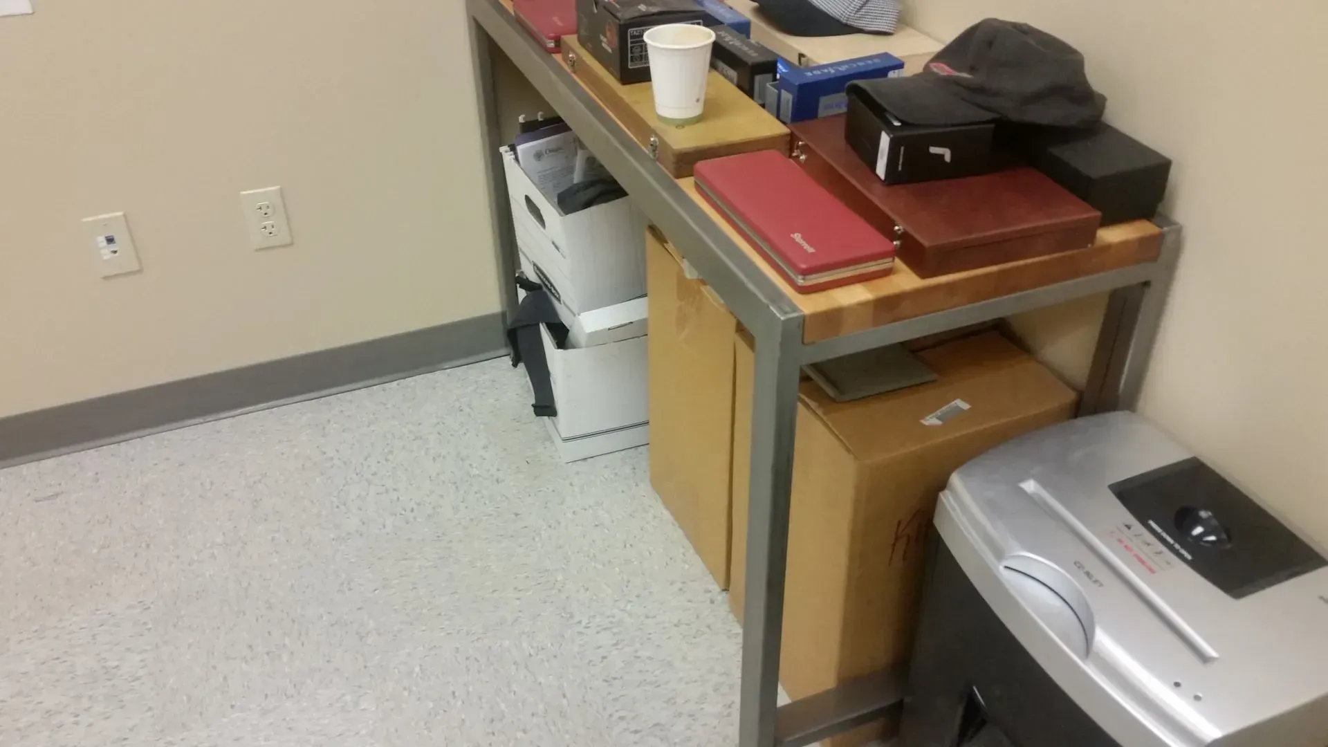 A desk in a corner holds office supplies: boxes, shredder, trash bin, and a cap. Light beige walls.