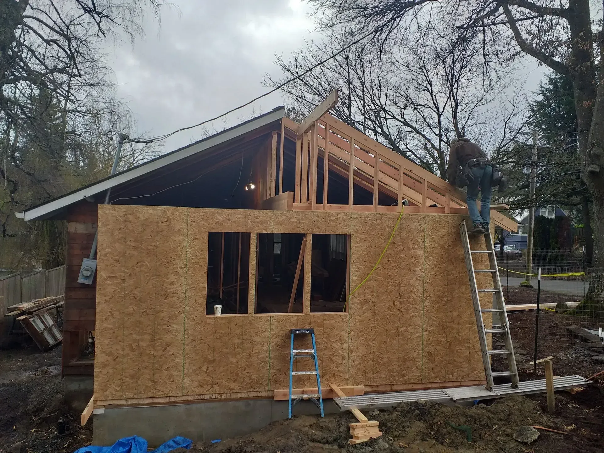 Construction of a building with a man on a ladder, wooden frame, plywood walls, and roof trusses.