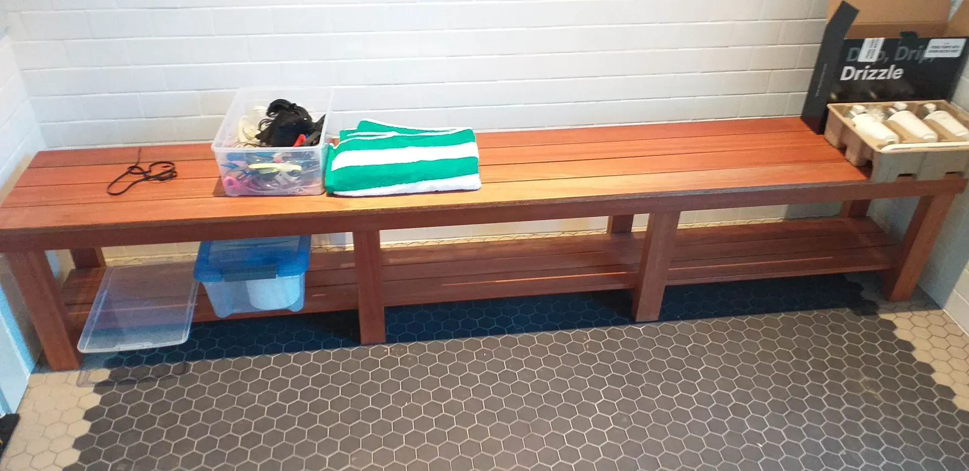 A wooden bench in a room with a patterned floor.  Items on the bench include storage containers and a green and white towel.