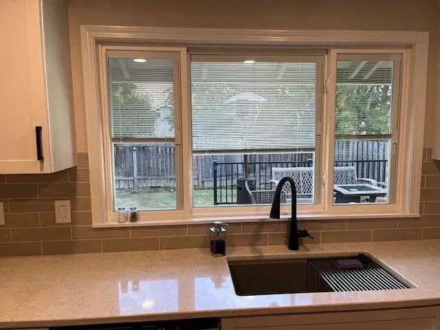 Kitchen sink under a window with a view of a backyard. Beige backsplash, white cabinets, and a black faucet.