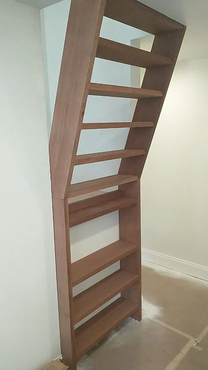 Wooden, tall bookshelf built into a wall, with slanted top and several shelves; tan and brown colors.