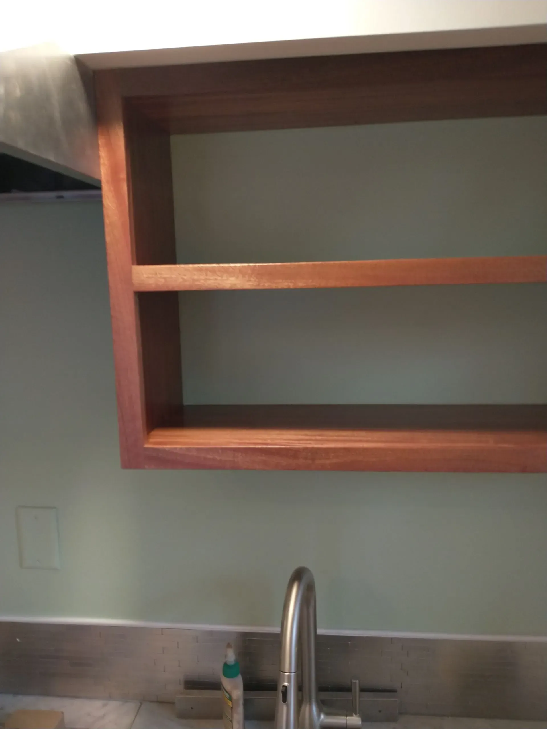 Wooden open shelf cabinet over a sink. Mahogany-toned wood against a light green wall.