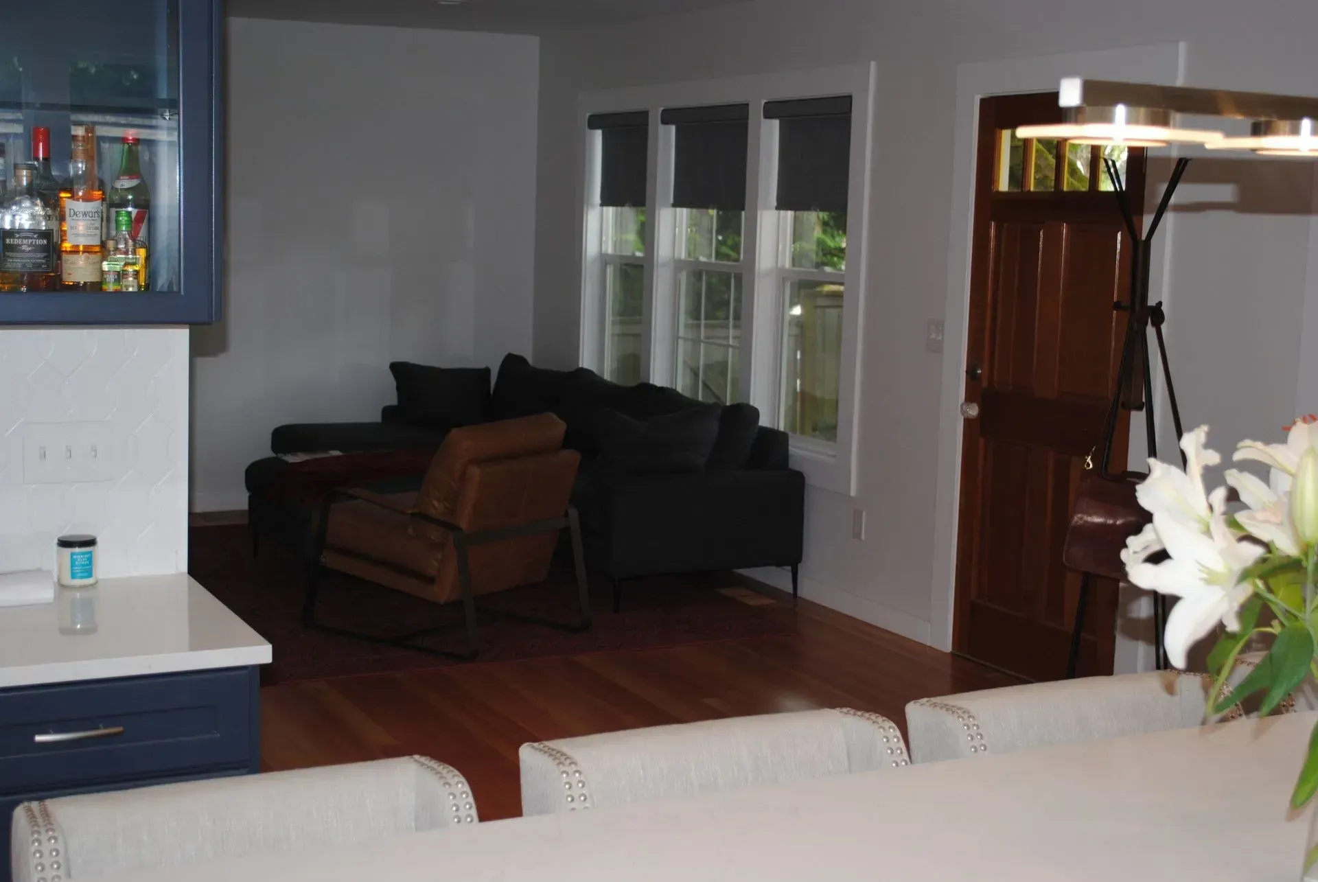 Living room with dark gray sectional, brown chair, wood floor, and window with black shades.