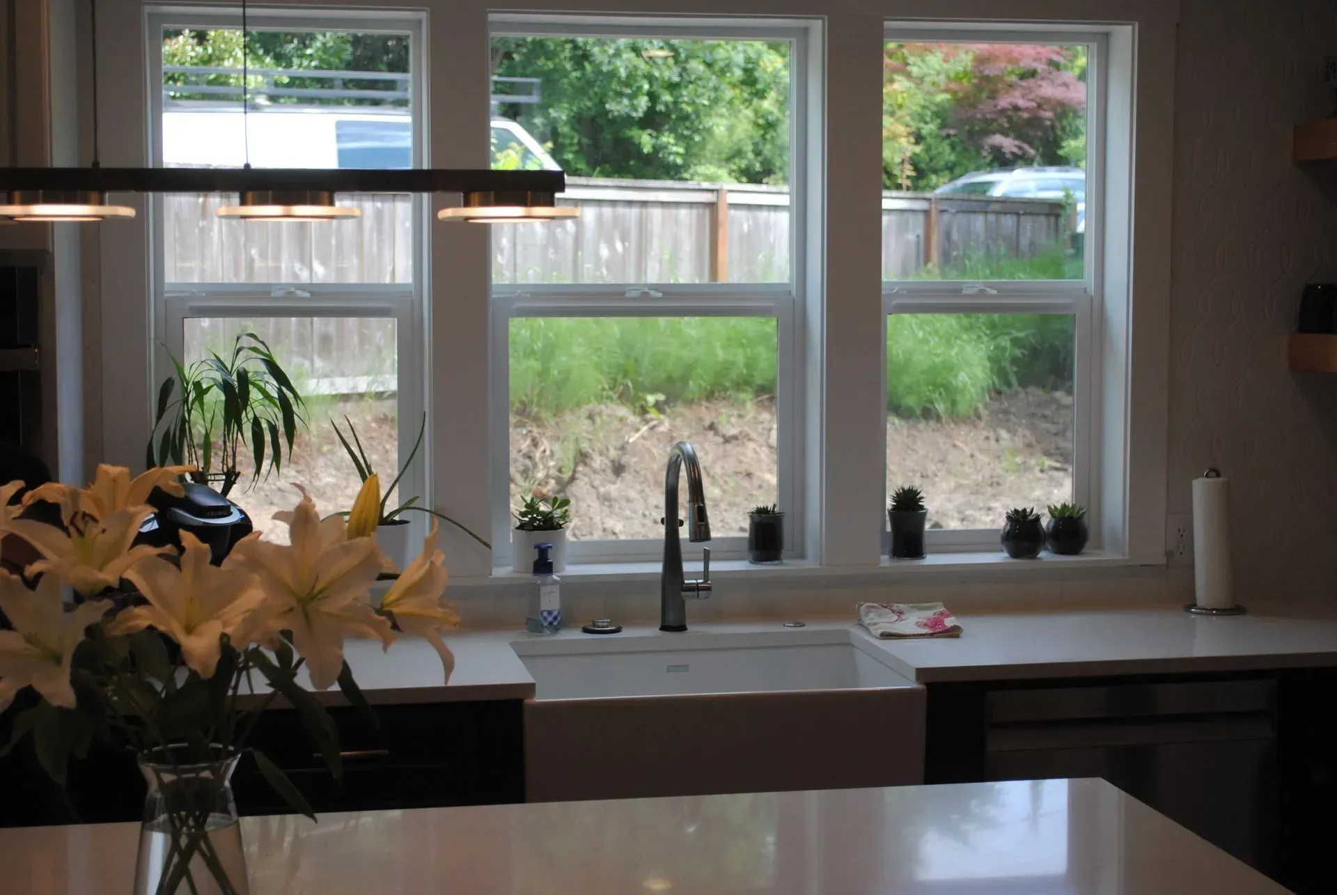 Kitchen with a white sink, counter, and windows overlooking a backyard with a wooden fence.
