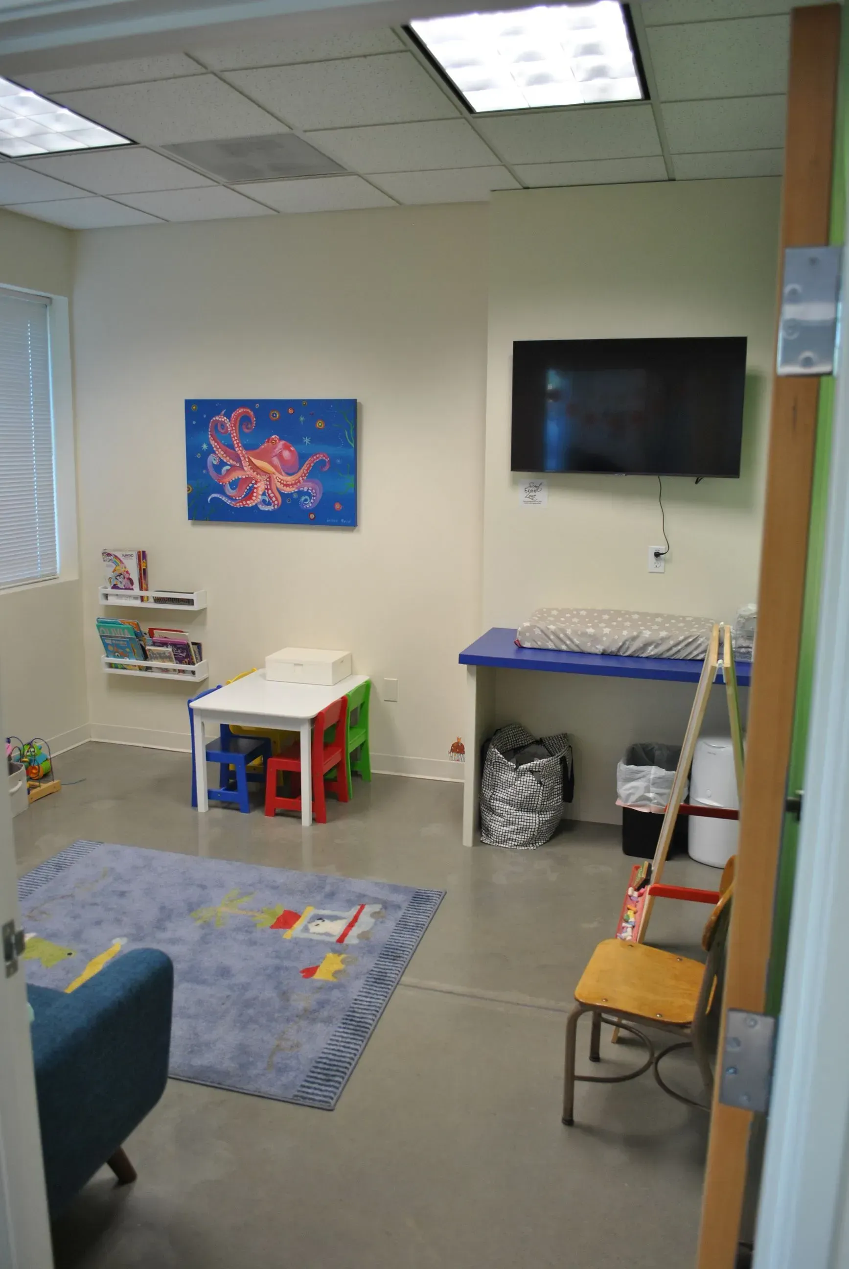 Child's playroom: Small table, toys, changing station, TV, octopus art, blue rug, and a partly visible blue armchair.