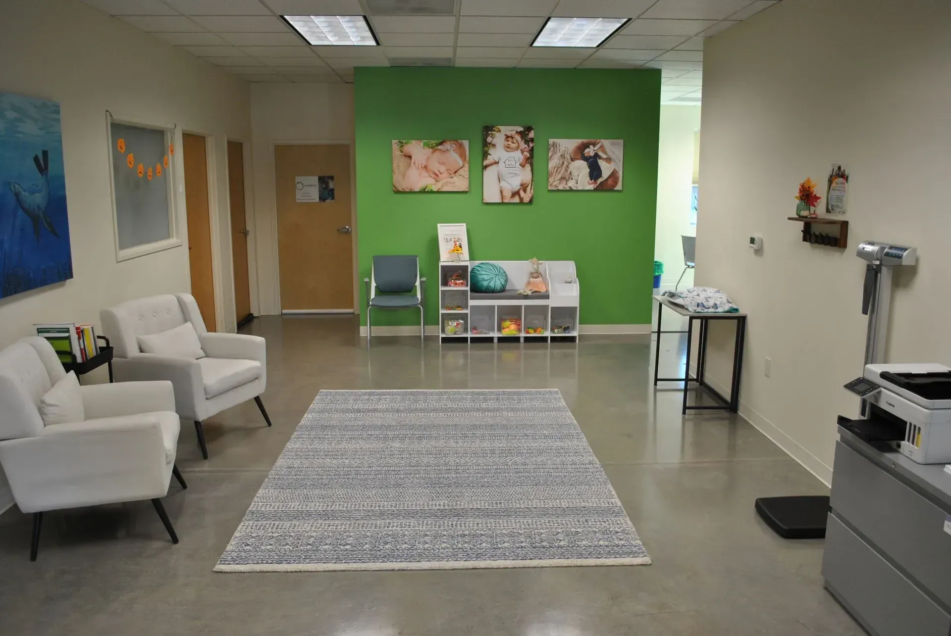Waiting room with white chairs, green wall with photos, rug, and office equipment.