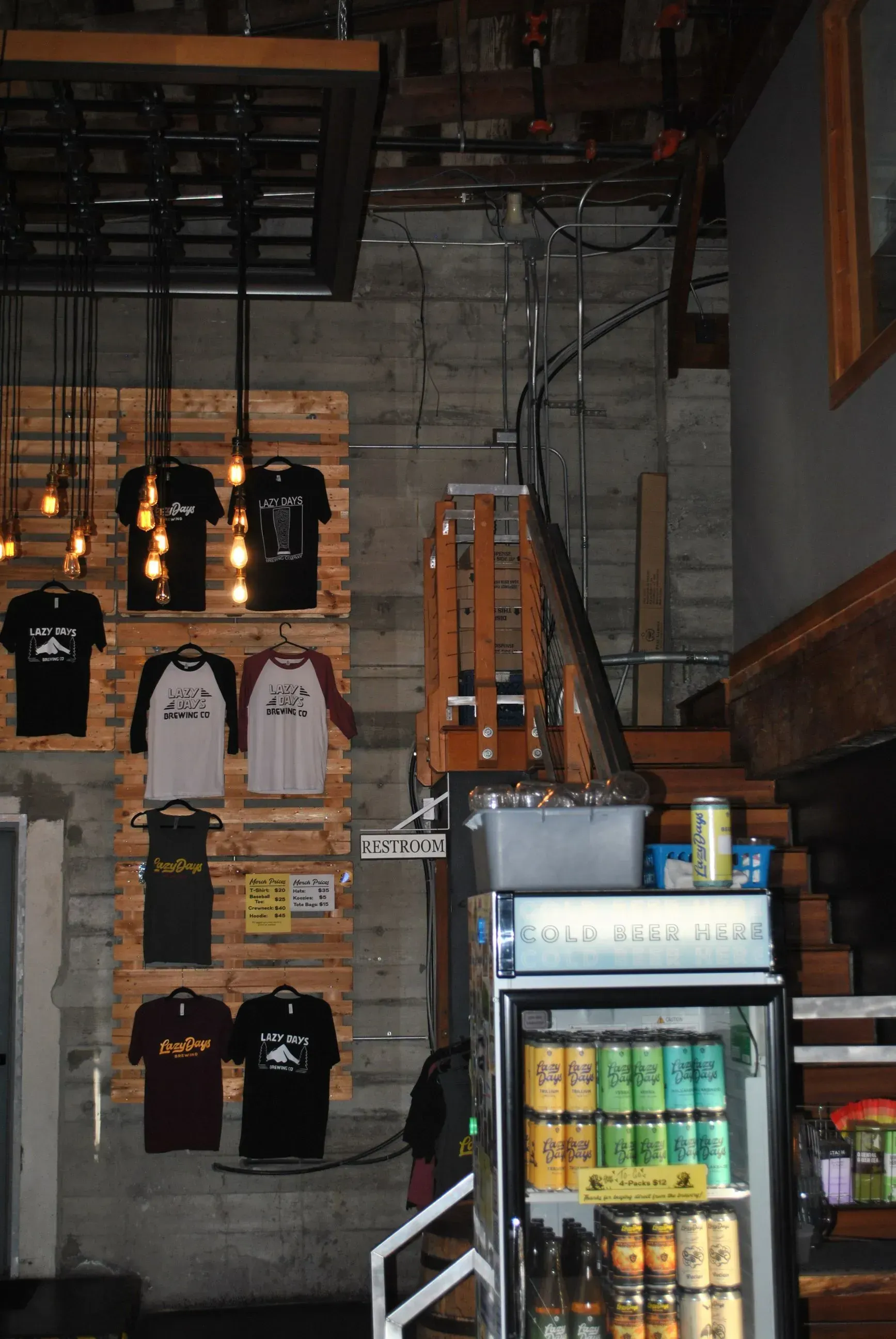 Inside a brewery: t-shirts on a pallet wall, glass-door fridge with drinks, stairs to a higher level, and exposed beams.