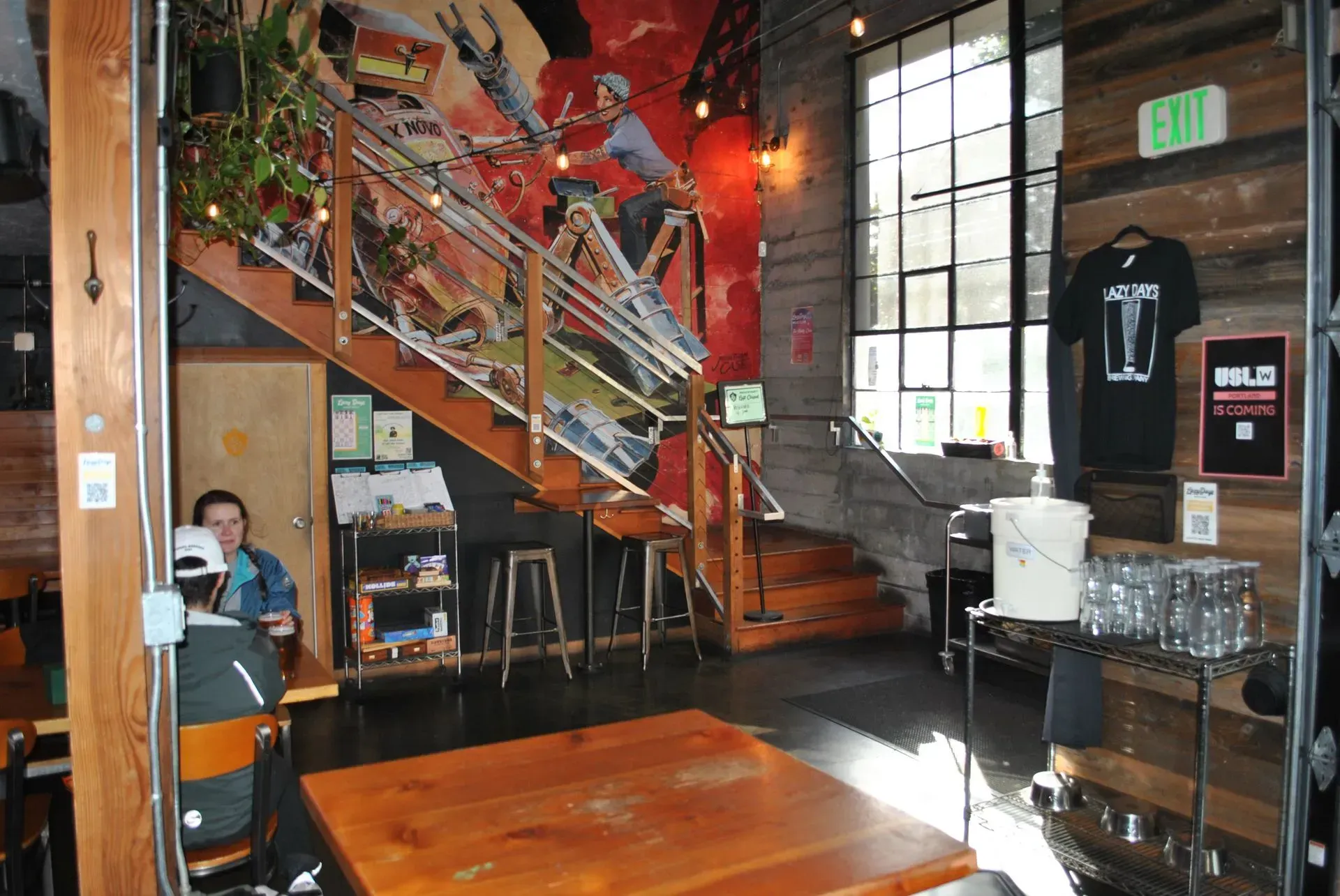 Interior of a cafe with a mural, staircase, and seating area. A person sits at a table.