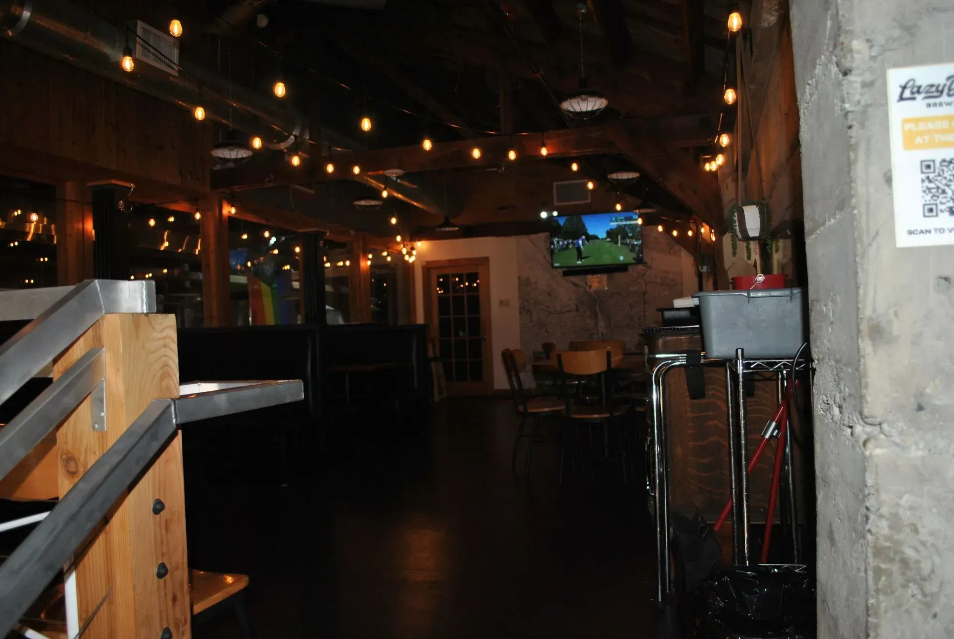 Interior of a bar or event space, with exposed ceiling, string lights, and a TV screen. Dark floor and neutral walls.