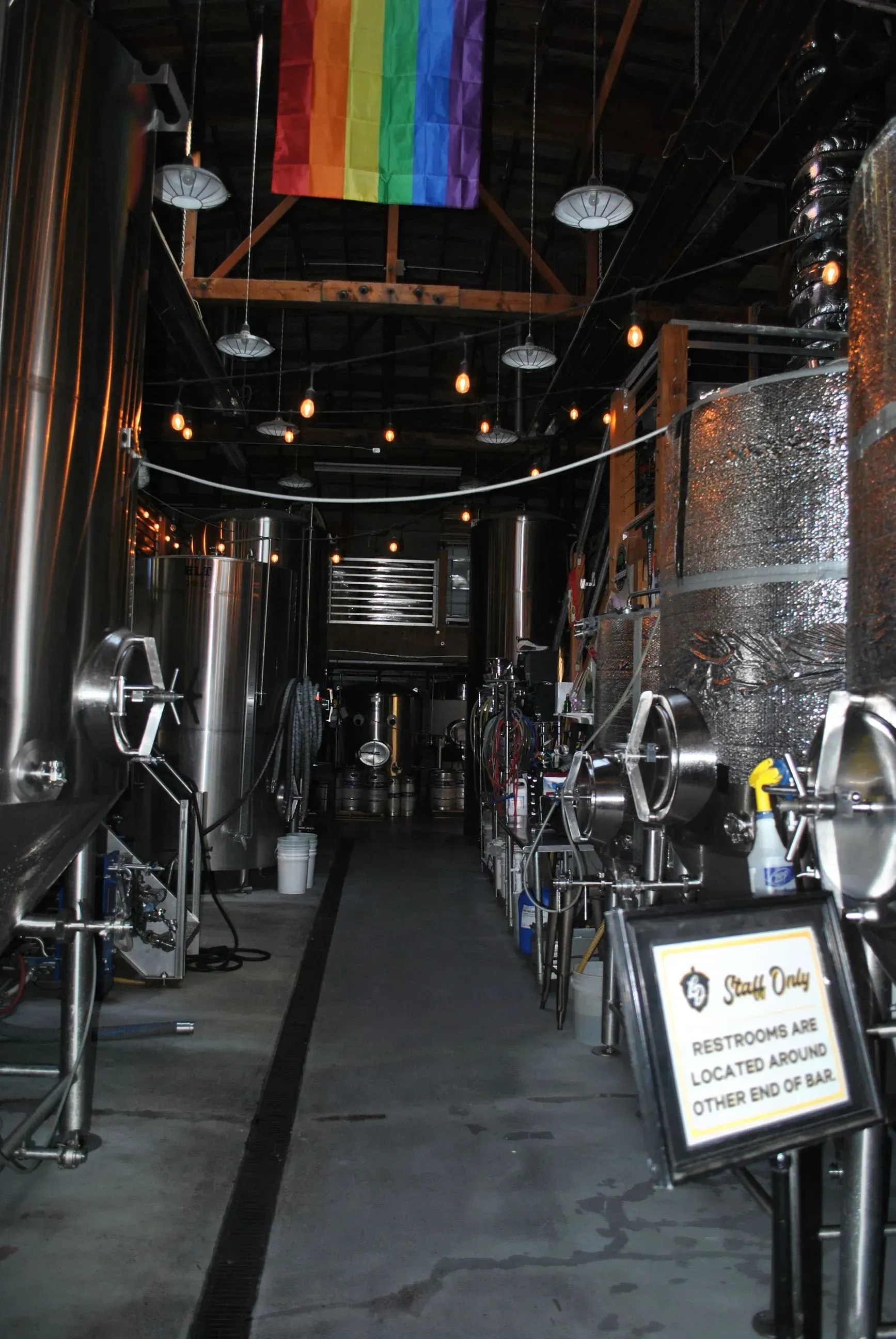 Inside a brewery, stainless steel tanks line a long hallway; a rainbow flag hangs overhead.