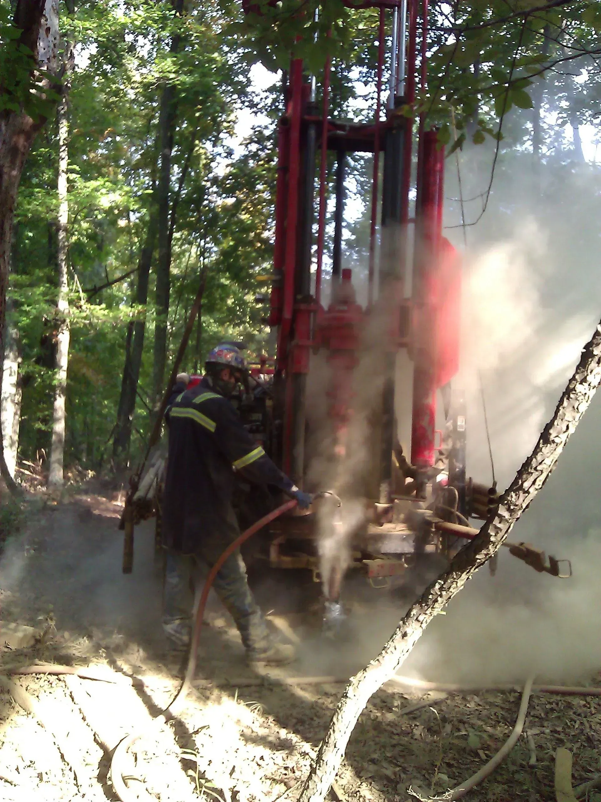 Person operating red drilling rig in a wooded area, dust and mist clouding the scene.