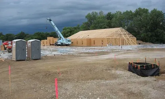 Construction site with wood-framed building, crane, and portable toilets; overcast sky.