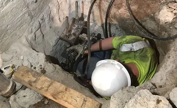 Worker in hard hat inside a hole, inspecting equipment. Yellow shirt, concrete environment.