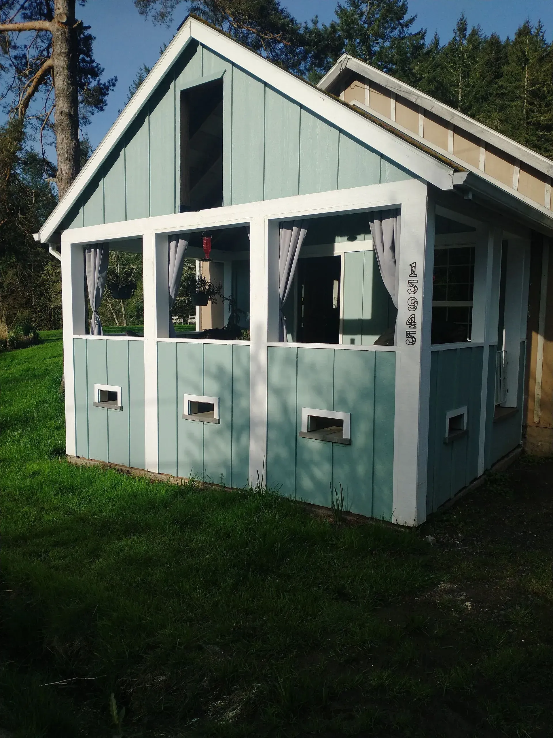 Small, light blue cabin with a porch. White trim, gray curtains. Set on green grass.