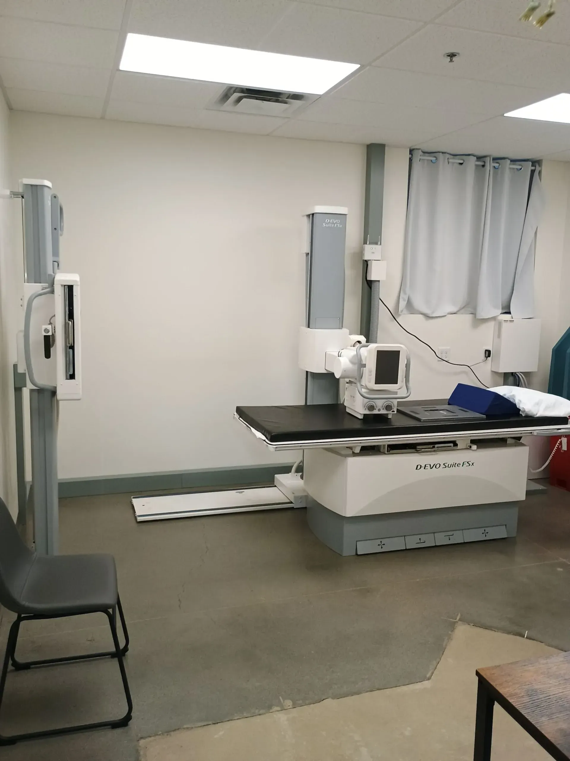 Medical examination room with X-ray equipment: table, stand, monitor, gray walls, and a black chair.