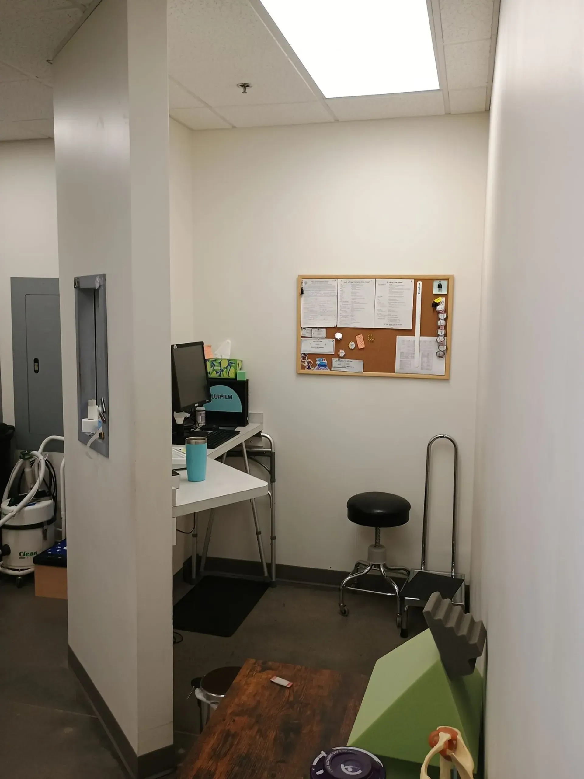 Office space with desk, corkboard, stool, and medical equipment. White walls and grey floor.