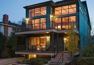 Modern blue house with large windows, balconies, and warm interior lighting at dusk.
