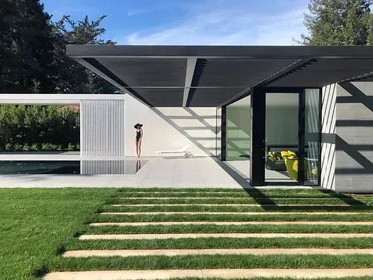 Modern home exterior with a shaded patio, pool, and green lawn. A person stands near the pool.