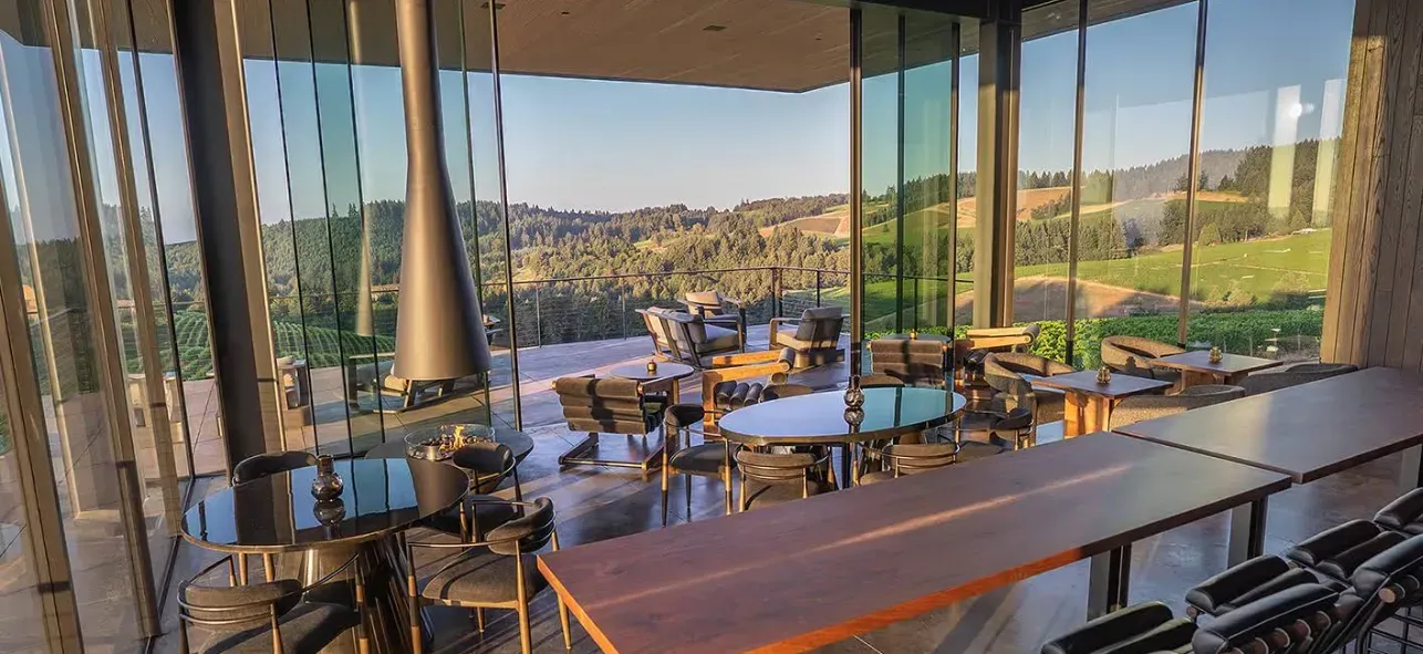 Modern dining area with floor-to-ceiling windows overlooking a hilly landscape. Features tables, chairs, and a fireplace.