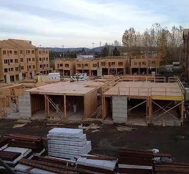 Construction site with wood-framed buildings in progress; materials and equipment visible. Cloudy sky.