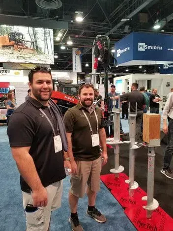 Two men stand at a trade show booth, near ground screw displays, smiles visible.