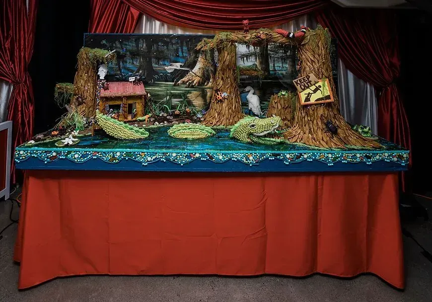 Diorama of a swamp scene with a house, trees, water, and signs, displayed on a table with a red skirt.