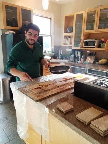 Man cooking in a kitchen, holding a pan. He stands by a countertop with wooden planks and a stovetop.
