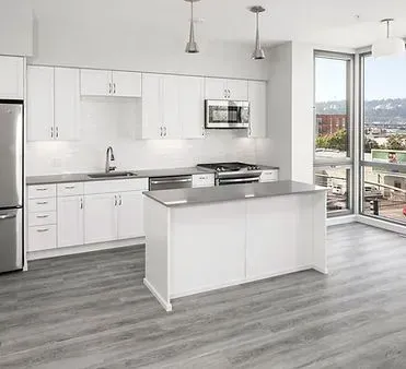 Modern white kitchen with stainless steel appliances, island, and large windows with cityscape view.