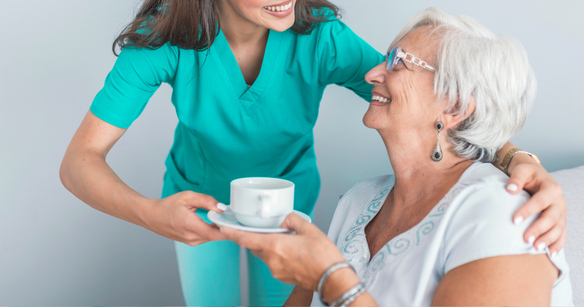 Woman in white scrubs assists an elderly woman in a wheelchair outdoors, smiling.