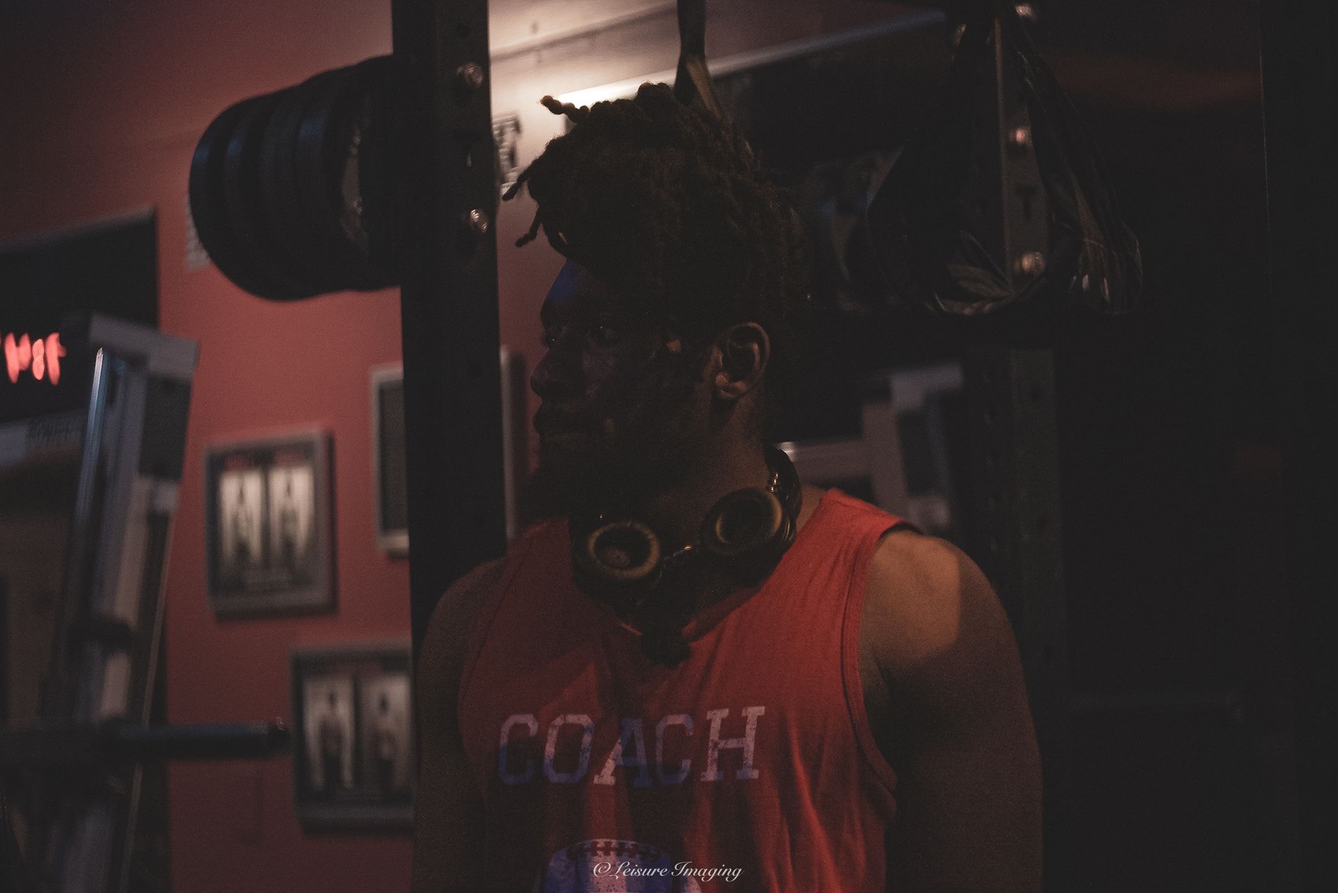 Man in a white tank top takes a gym selfie, showcasing arm muscles. Gym setting with weights in background.