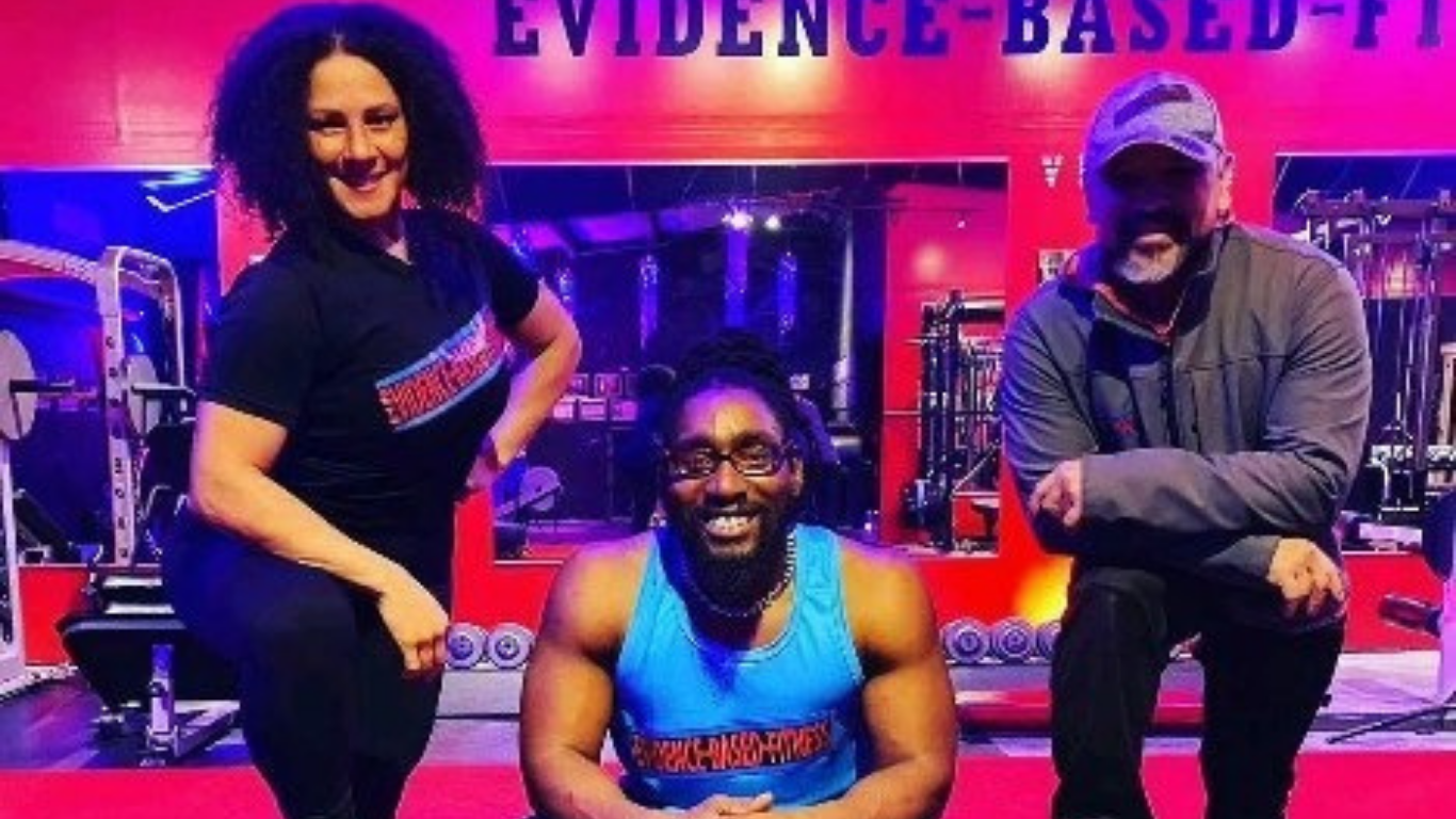 Three people smiling in a gym: woman flexing, man kneeling, and another man smiling. 