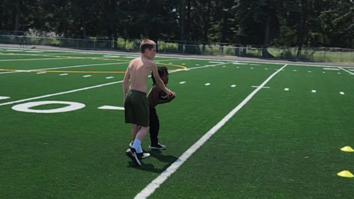 Two people on a football field practice holding the football. One person is shirtless.