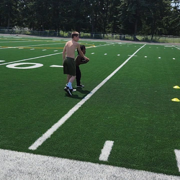 Two people on a green turf field, one shirtless, appear to be practicing football.