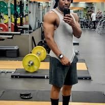 Man in a tank top and shorts, flexing in a gym.  Weight plates and equipment visible.