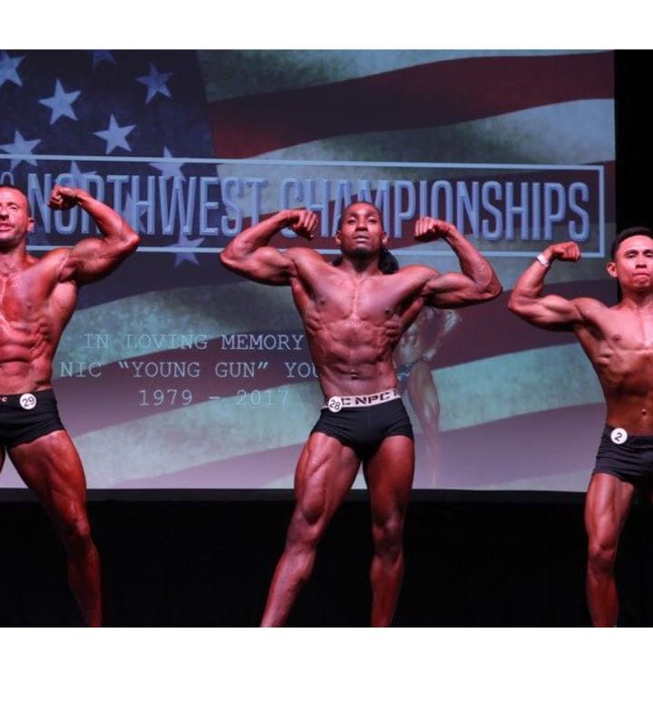 Three bodybuilders flexing onstage at the Northwest Championships. They are wearing black trunks. American flag background.