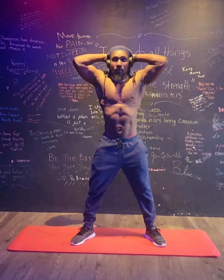 Man with hands behind head, in athletic pose on mat, in front of graffiti wall.