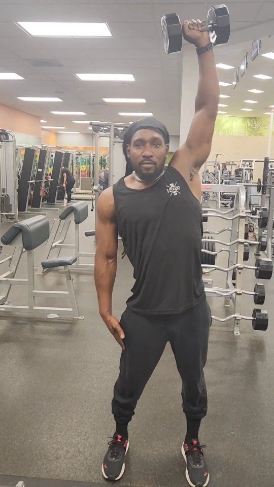 Man in black workout clothes lifting a dumbbell overhead in a gym.