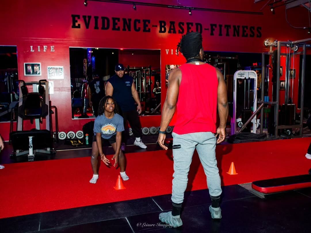People exercising in a gym with red floor and walls. A trainer observes a person holding weights, with others 