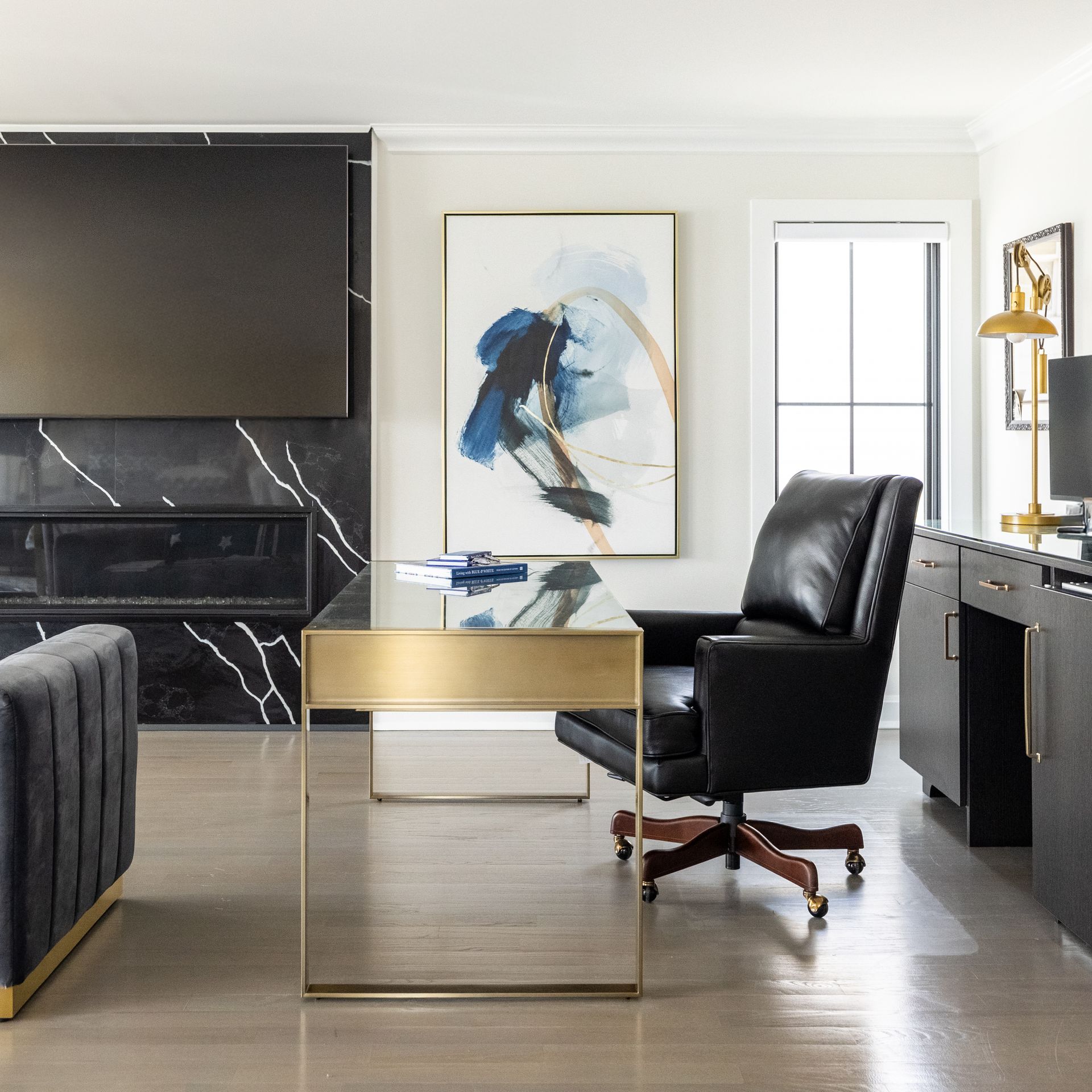 Elegant home office with a black leather chair and a gold-trimmed glass desk. Artwork hangs on a cream wall.