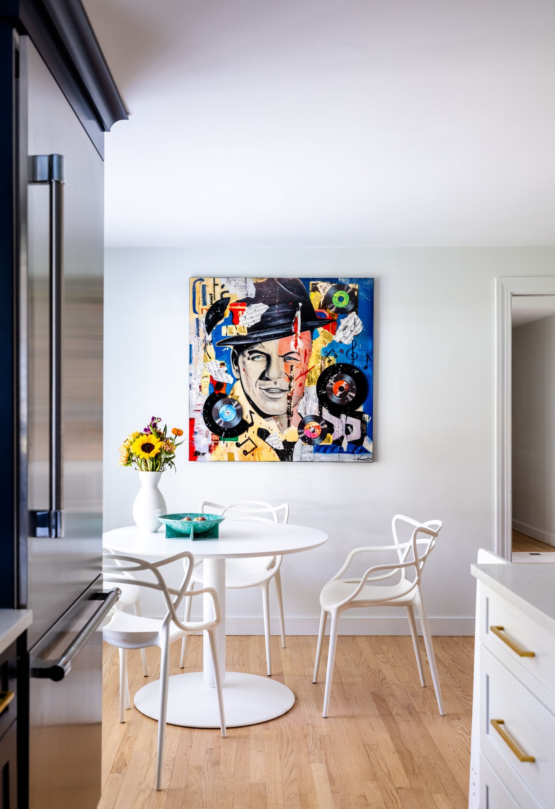 Dining area with white table and chairs, colorful artwork, sunflowers, and light wood flooring.